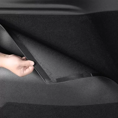 Hand pulling a strap to lift a floor panel inside a car trunk to access under-floor storage in the mat area.