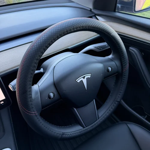 Tesla steering wheel wrapped in black perforated leather with red stitching, inside a modern car cabin.