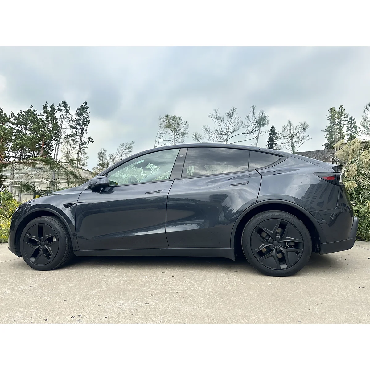 Side profile of a dark gray Tesla Model Y parked on a driveway with trees in the background.