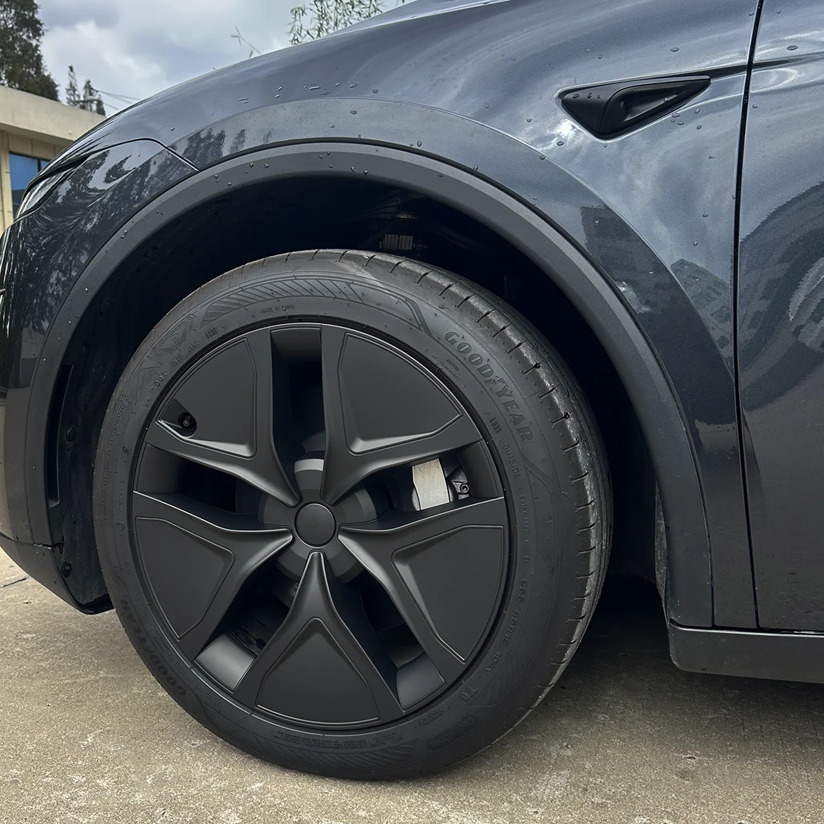 Close-up of a dark gray car's front left wheel: matte black alloy rim, Goodyear tire, and visible brake caliper, with raindrops on the body.